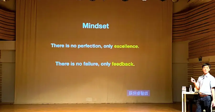 Dr. Su lecturing beside a projected slide reading: There is no perfection, only excellence. There is no failure, only feedback.